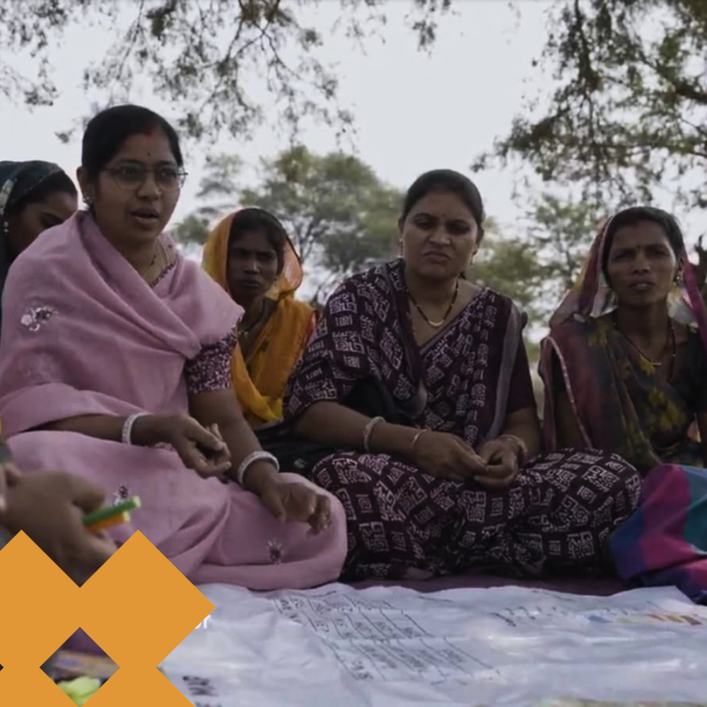 Group of Indian women sitting on a white sheet outdoors, engaged in discussion or activity together.