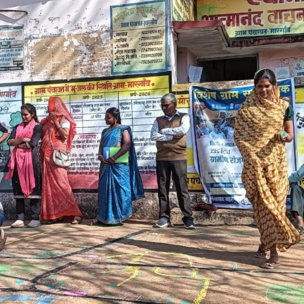 A row of villagers, mostly women in colorful sarees, stand in front of a wall covered with community posters and banners; chalk drawings are on the ground in front of them, suggesting a local event or gathering.