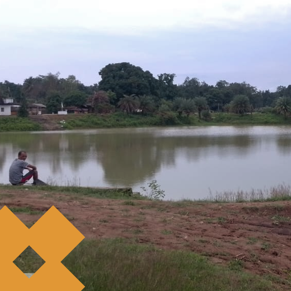Person sits on grassy bank by a calm lake, looking toward the water; rural houses and trees on the far shore; orange watermark in bottom-left corner.