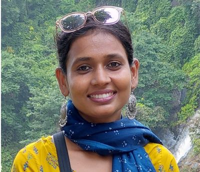 Smiling woman wearing a yellow top and blue scarf, large earrings, sunglasses on her head, outdoors with greenery in the background.
