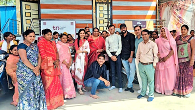 Group of men and women in colorful traditional dress posing for a group photo outside a bright striped building and banners in the background.