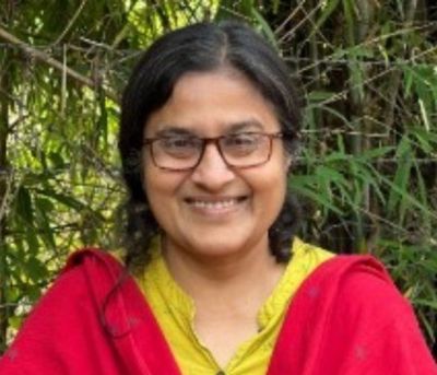 Smiling woman with glasses wearing a red shawl over a yellow top, standing in greenery behind her.