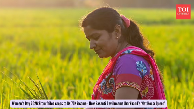 Woman in a purple outfit with a pink scarf stands in a sunlit golden crop field, looking down.
