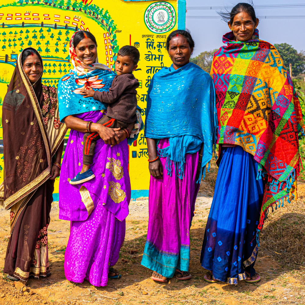 Group of five Indian women in colorful saris pose outdoors beside a bright yellow mural, with a small child held by one woman.