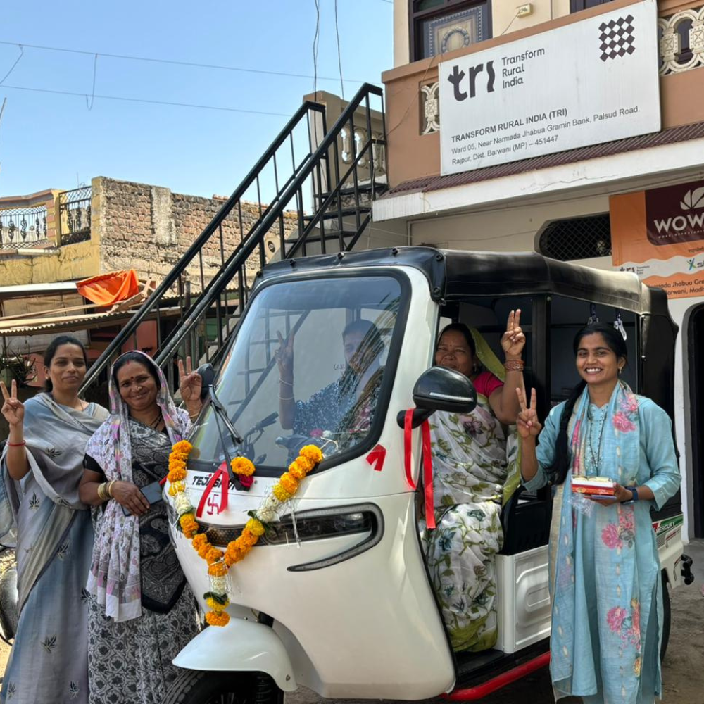 Smiling group of Indian women around a white auto-rickshaw decorated with marigold garlands, giving peace signs.