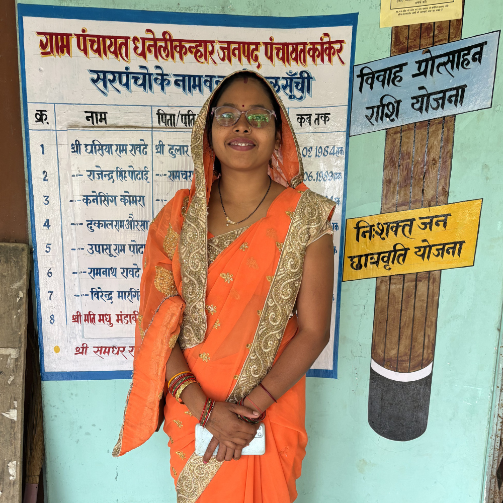 Young woman in a bright orange sari with gold trim, wearing glasses and bangles, smiles while standing in front of a blue community notice board with Hindi text.