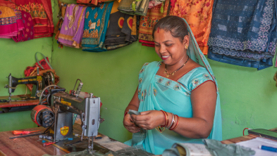 Woman in a turquoise sari sits at a sewing machine, stitching fabric in a textile shop with colorful rolls hung on a green wall behind her.