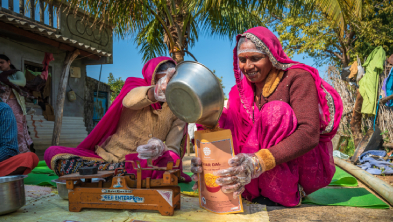 Two Indian women in bright pink and red traditional clothes sit on a mat outdoors, one pouring from a metal pot into a container while the other holds a labeled box nearby. Palm trees and a small shop backdrop the scene, suggesting an outdoor market or roadside stall.