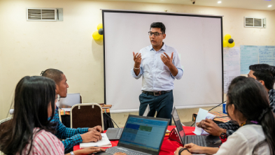 Teacher standing in front of a projection screen, teaching students around a table with laptops.