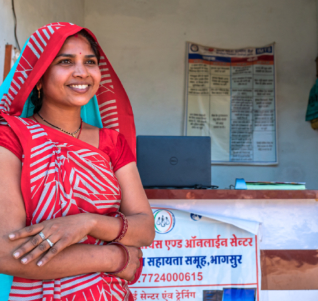 Smiling Indian woman in a red and white sari stands with arms crossed in front of a desk with a laptop at a learning center.