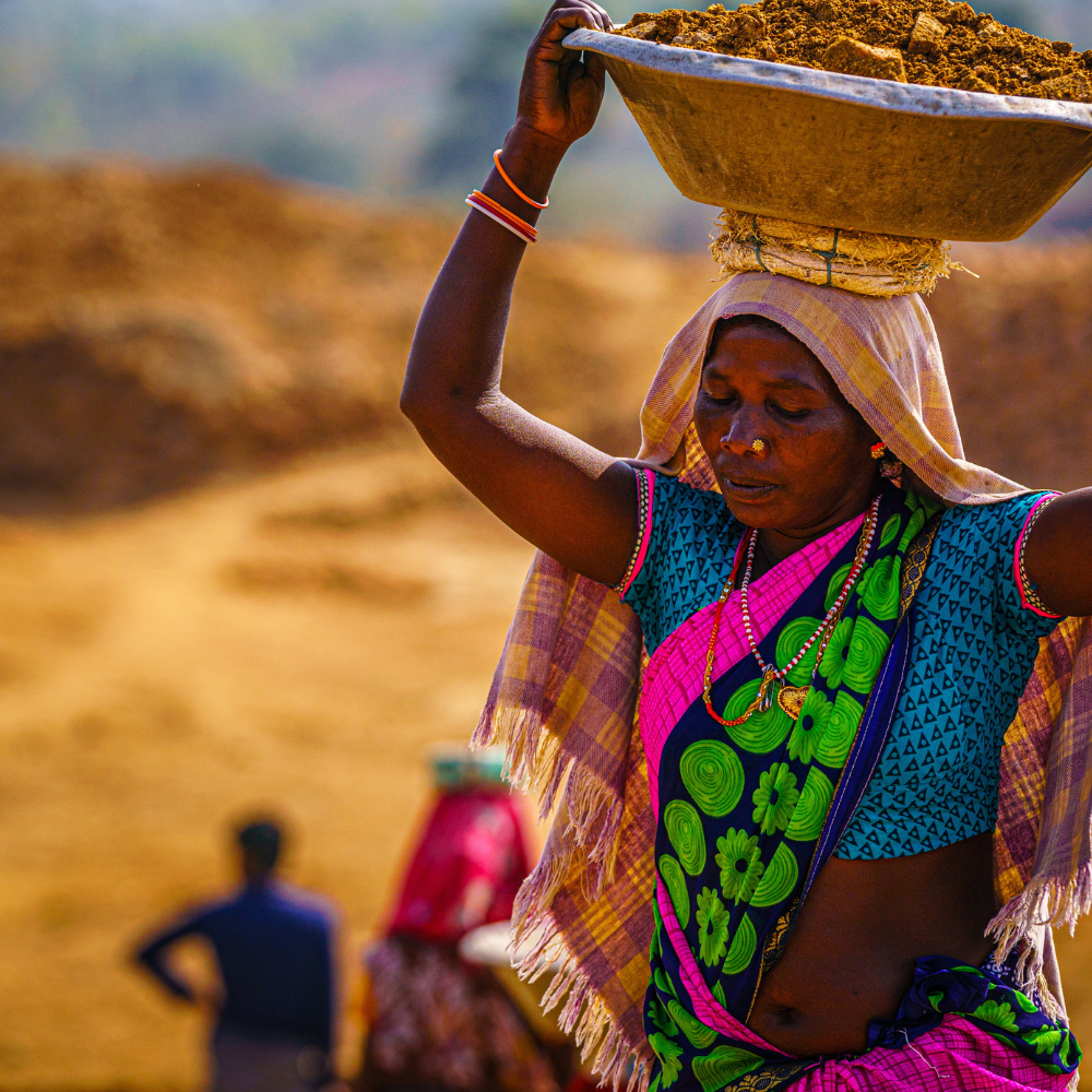 Woman in colorful clothing balances a large basket filled with soil on her head in a dusty outdoor setting.