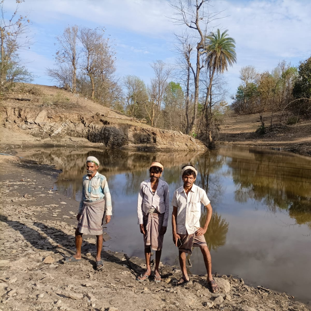 Three men in traditional clothing stand on a rocky riverside with a calm river reflecting trees and a blue sky in the background.