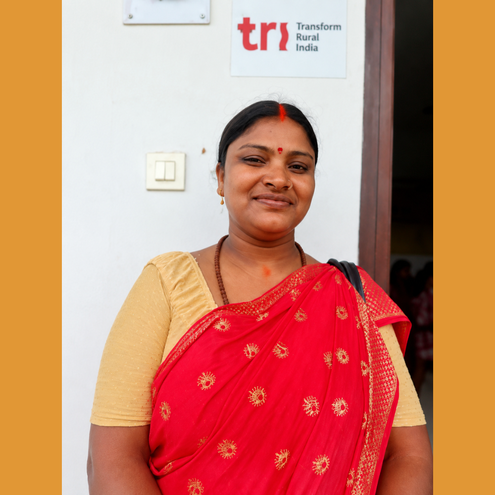 Smiling woman in a red sari stands in front of a wall with a TR Transform Rural India sign; warm orange borders frame the scene.