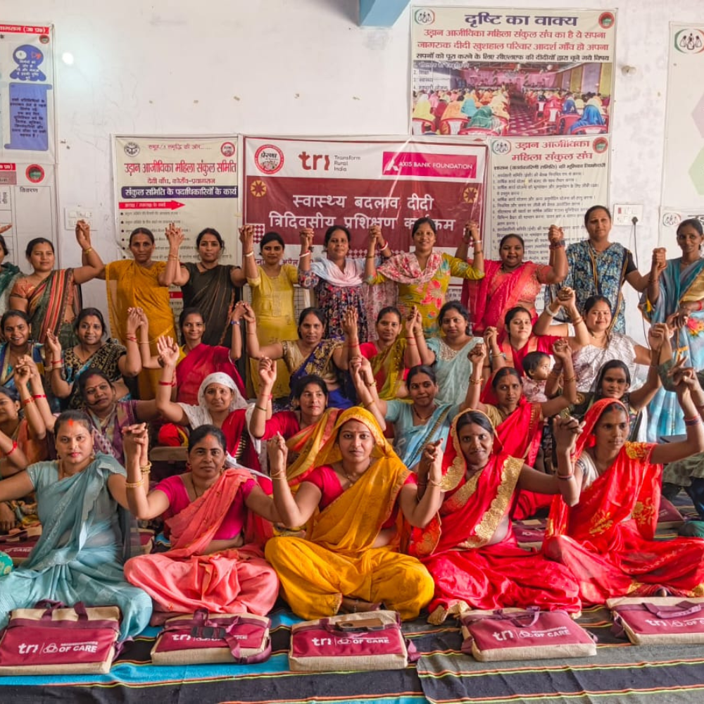 Group of Indian women in colorful sarees posing in a hall during a community program, many raising their hands in unison, with banners and bags from sponsors in the background.