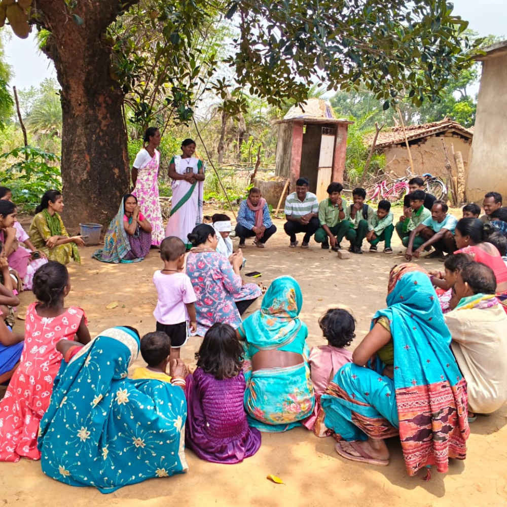 A diverse group of villagers sits in a circle under a large tree, outdoors in a rural village setting.