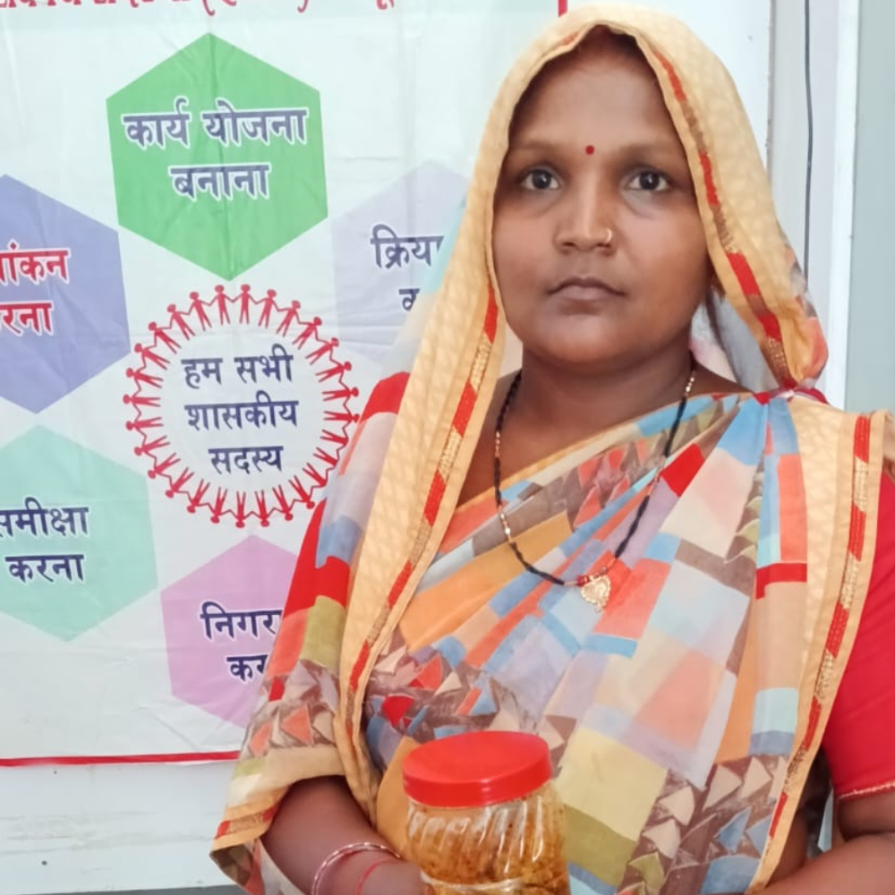 Indian woman in a colorful saree with a red bindi, holding a jar of orange food in front of a program banner.