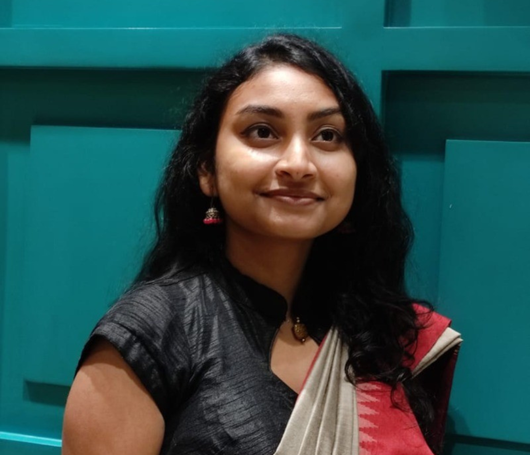 A woman with long dark hair wearing a dark blouse and a red-beige sari stands against a teal panel background, smiling softly.