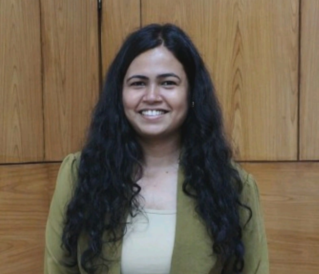 Smiling woman with long dark curly hair standing in front of a wood-paneled wall, wearing a green blazer and light top.