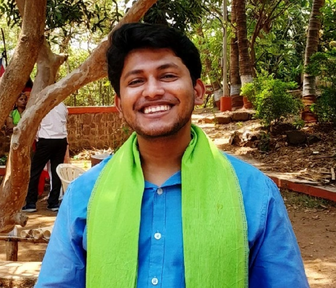 Young man smiling outdoors, wearing a blue shirt and bright green scarf in a sunny garden setting.